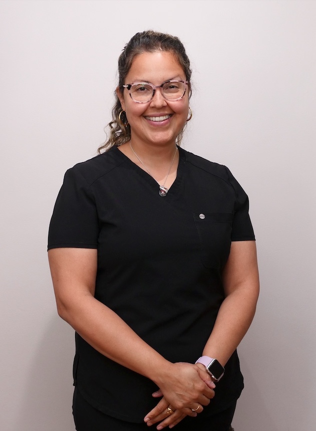 The image shows a person, likely a healthcare professional, wearing scrubs and standing against a plain background. They are smiling and appear to be posing for the photograph.