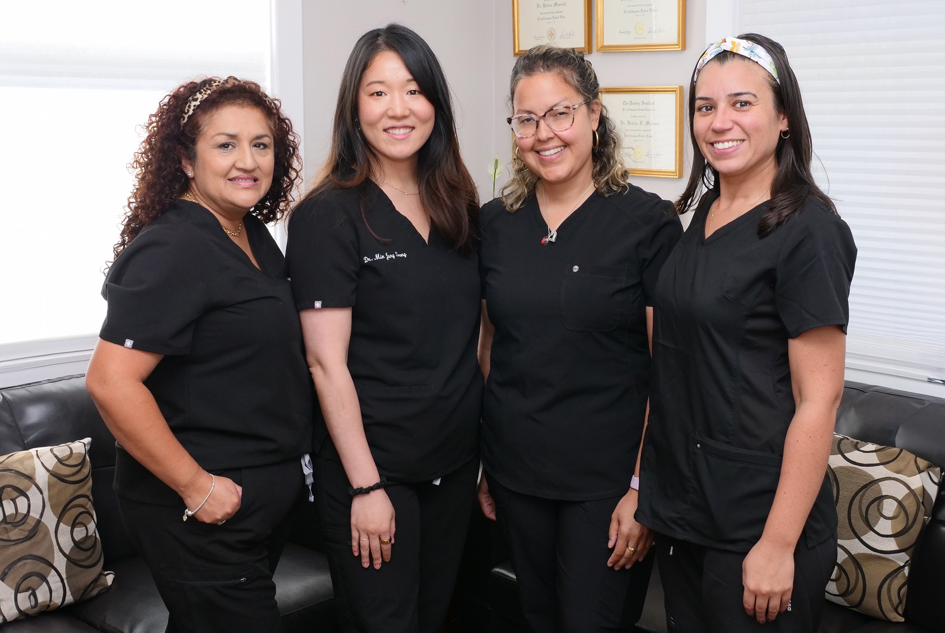 The image is a photograph of four individuals, likely nurses or healthcare professionals, posing for the camera in an office setting. They are standing together, smiling and appear to be dressed in uniforms that include white coats, stethoscopes, and name tags.