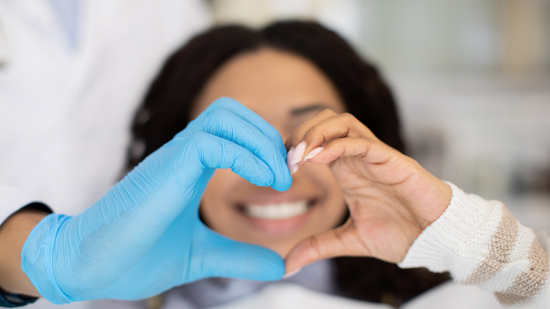 A medical professional in a blue gown and gloves holds up a heart-shaped object, symbolizing care or love, while a patient with bandages on their face smiles at the camera.