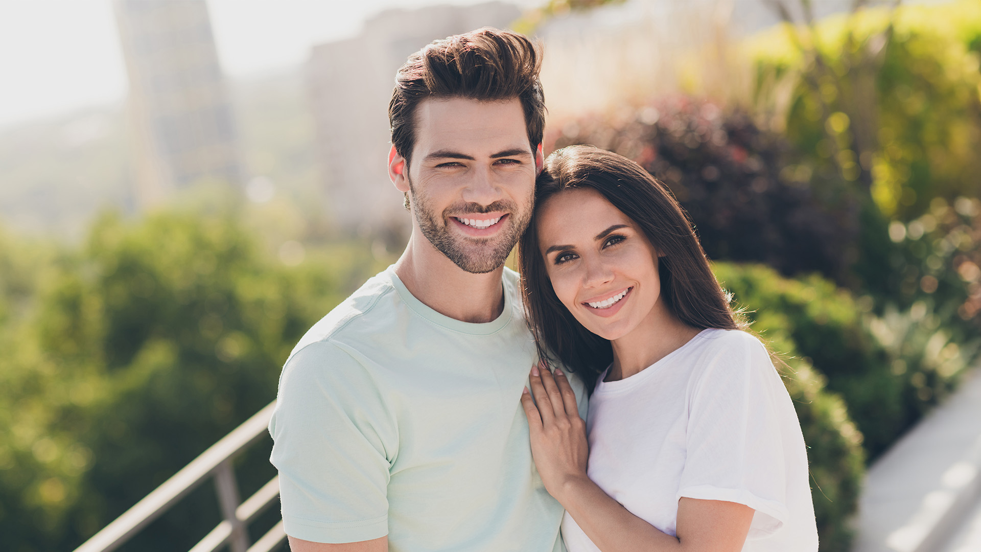 This is a color photograph of two adults, a man and a woman, posing for the camera with smiles. They are outdoors during daylight hours, standing close together, with the man on the left wearing a light blue t-shirt and dark pants, and the woman on the right in a white sleeveless top and dark pants.