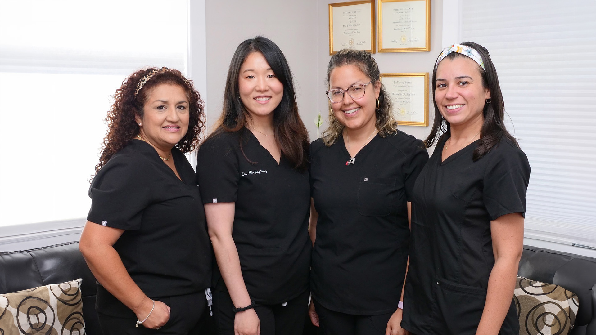 The image shows four individuals posing for a photograph, likely in a professional setting. They are wearing scrubs and standing side by side with smiles on their faces.