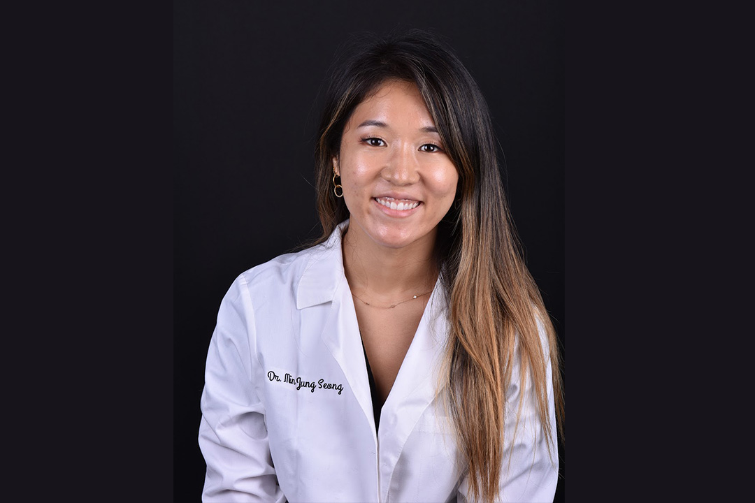 A smiling woman wearing a white lab coat, standing against a dark background.