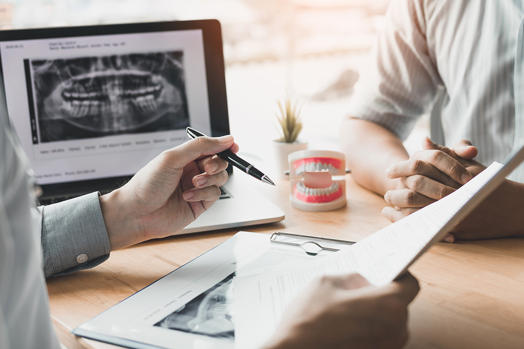 In the image, a man and woman are seated at a desk with a laptop, a tablet displaying dental X-rays, and a pen. The man is pointing to something on the tablet while the woman appears to be taking notes or reviewing documents.
