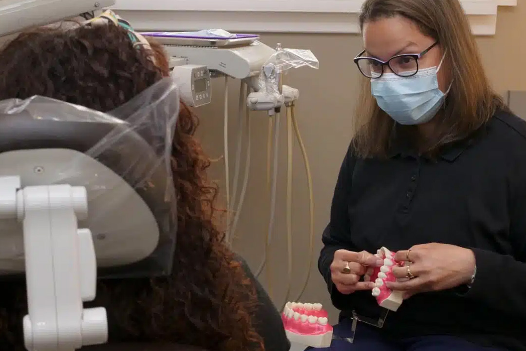 A woman in a dental office, wearing a surgical mask and glasses, is working on a patient s teeth.