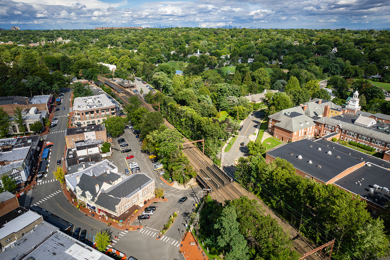 Aerial view of a suburban neighborhood with tree-lined streets, parked cars, and a mix of residential and commercial buildings.
