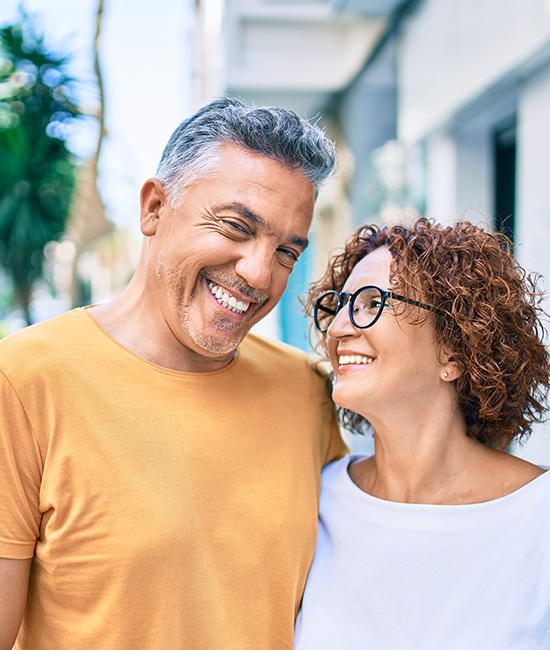 The image shows a man and a woman, both smiling, embracing each other in front of a building.
