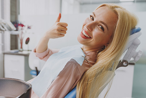 A woman with blonde hair is sitting in a dental chair, giving a thumbs-up gesture while wearing a face mask.