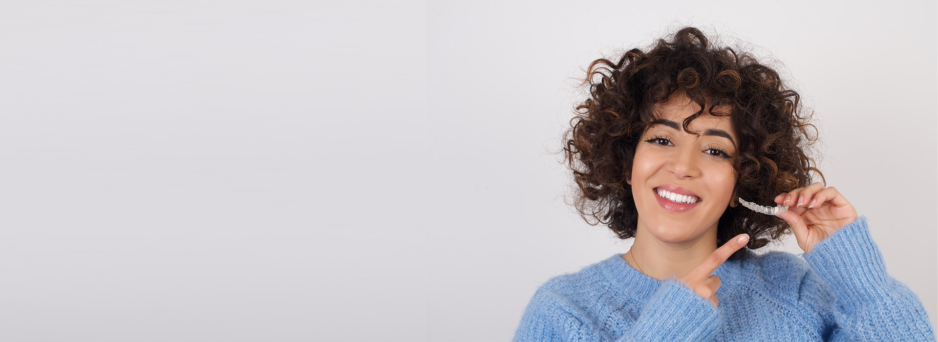 A woman with curly hair is smiling and holding a phone to her ear, set against a plain background.