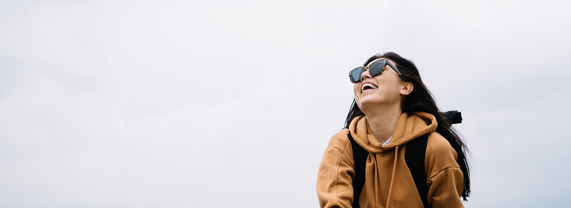 A person in a tan jacket and sunglasses, smiling and looking upwards while standing outdoors.