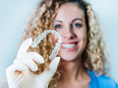 The image shows a person holding up a clear dental retainer, which is a type of orthodontic appliance used to maintain the alignment of teeth after braces are removed.