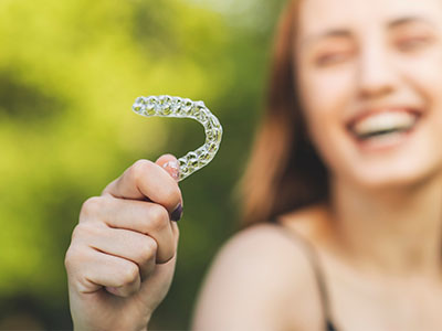A young woman is smiling and holding a transparent dental retainer between her fingers.