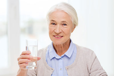 The image shows an elderly woman holding a glass of water, smiling slightly, and looking directly at the camera.