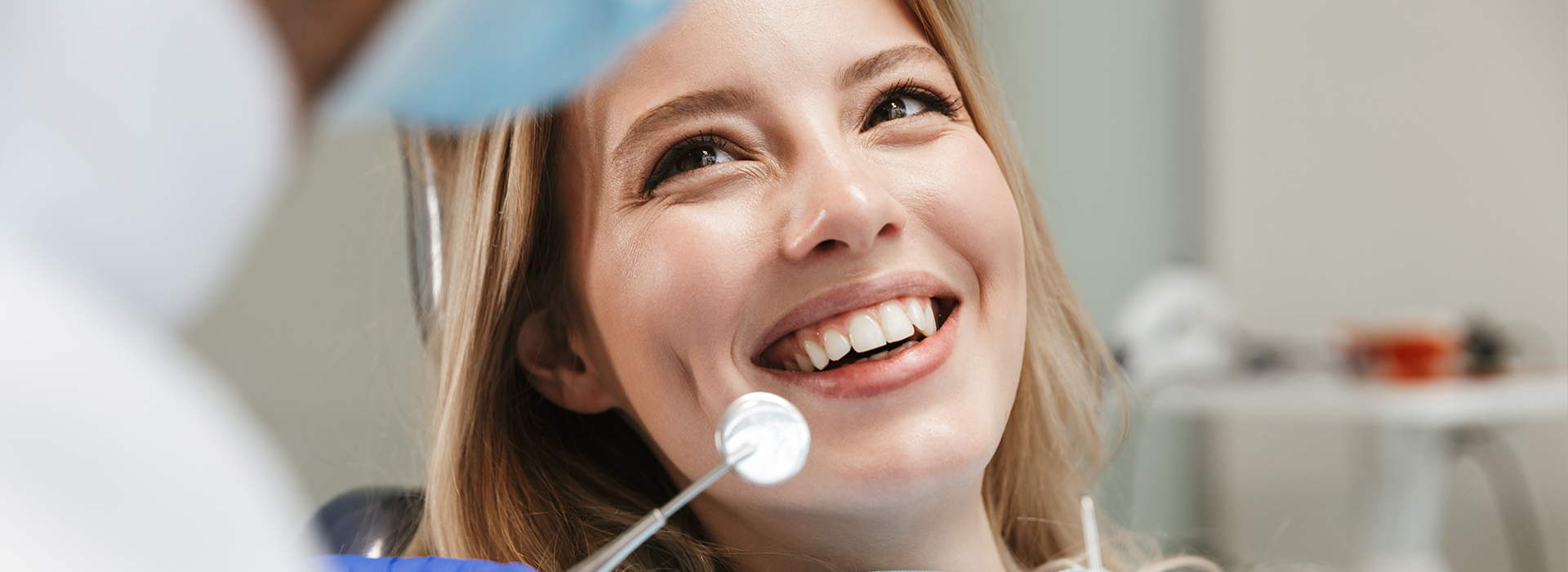 A young woman with a broad smile is seated in front of a dental professional, who seems to be examining her teeth while she looks directly at the camera.