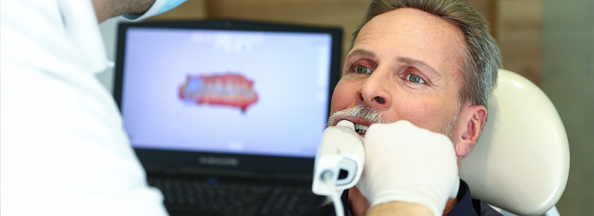 In the image, a man is seated in a dental chair, receiving dental care from a professional who appears to be using a computer or digital device for consultation.