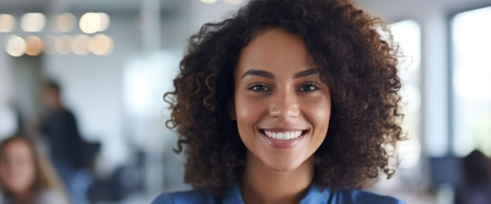 A smiling woman with curly hair, wearing a blue top, stands in front of an office background.