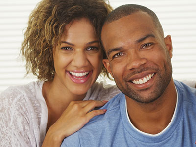 The image shows a man and woman smiling at the camera, with the man wearing a short beard and the woman with curly hair. They appear to be in a happy, intimate pose, possibly a couple.