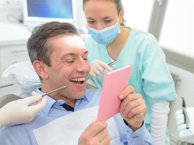 The image shows a dentist, possibly male, sitting in front of a pink card while holding it up with his right hand. He is smiling and appears to be looking at the camera. Behind him, there are two individuals wearing medical scrubs and masks  one is seated on a chair with dental instruments, and the other stands behind them, also wearing a mask. They all seem to be in a dental office setting.