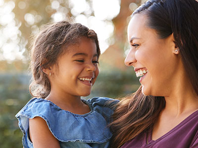 A smiling woman in a purple shirt holding a laughing child with brown hair, both outdoors.