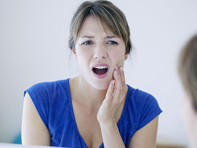 A woman with her mouth open, showing teeth and expression of concern or surprise, looking at her reflection in a mirror.