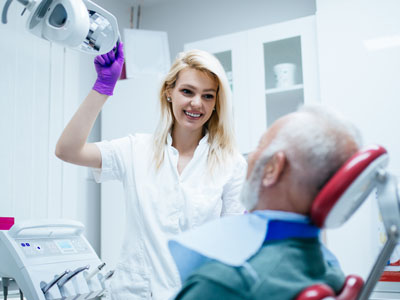 A woman in a white coat is assisting an elderly man with dental treatment, with the woman holding up a mirror to show the patient s teeth.