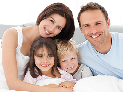 A family of four, including a man, woman, and two children, smiling together in a bedroom setting.