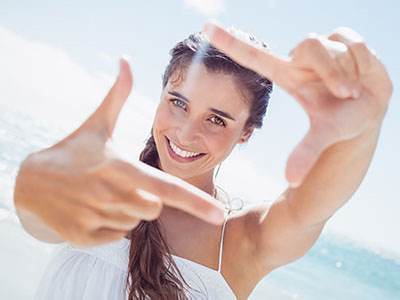 A woman is taking a selfie at the beach, smiling and pointing towards the camera.