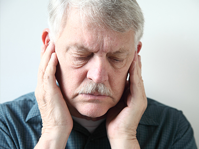 The image shows an older man with his eyes closed, holding his face with both hands. He appears to be in a state of discomfort or pain.